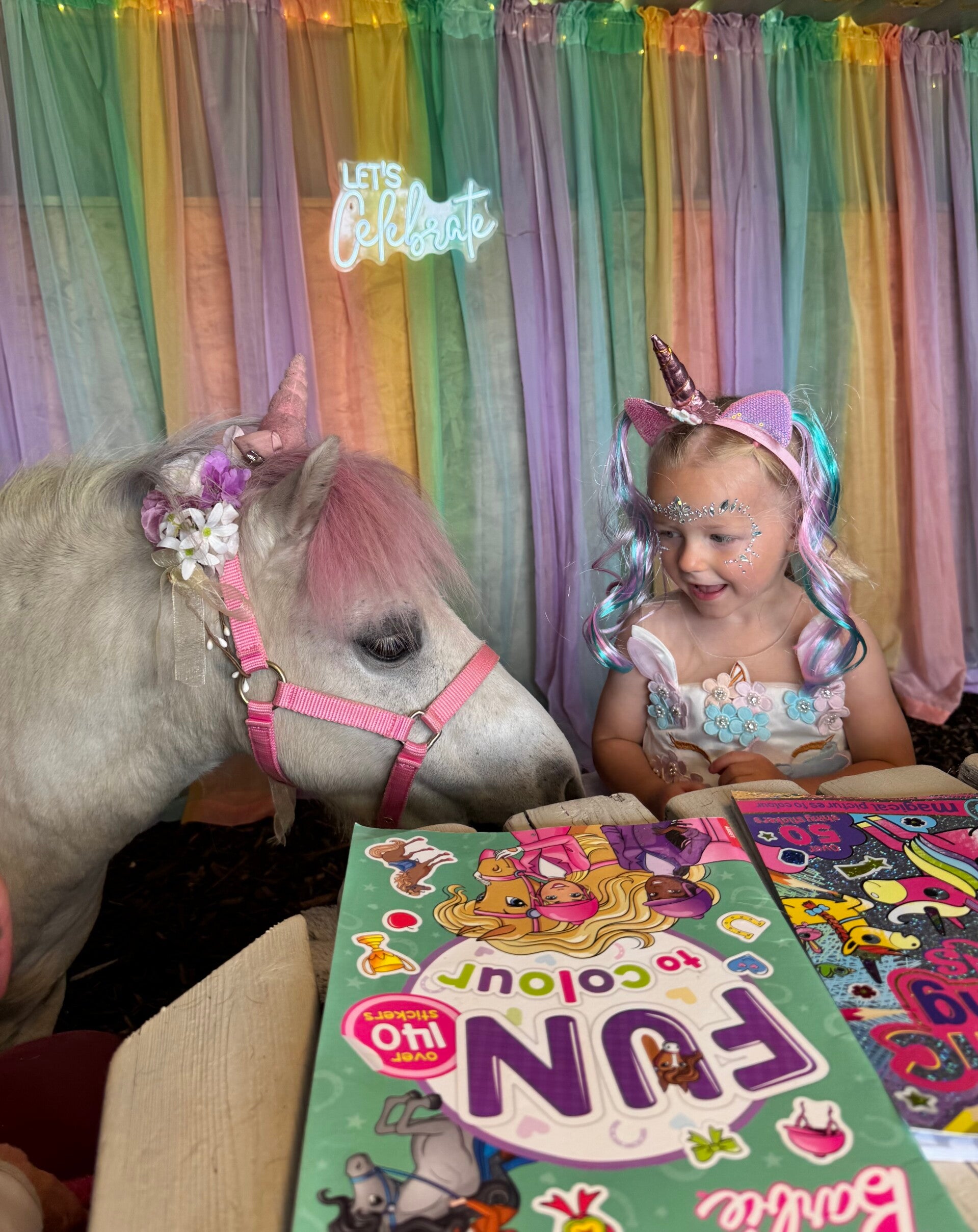 A happy child sat by the craft table with colouring books smiling at the white unicorn pony stood beside them. In the background there is a pastel coloured material backdrop with a neon 'let's celebrate' sign in the middle. 
