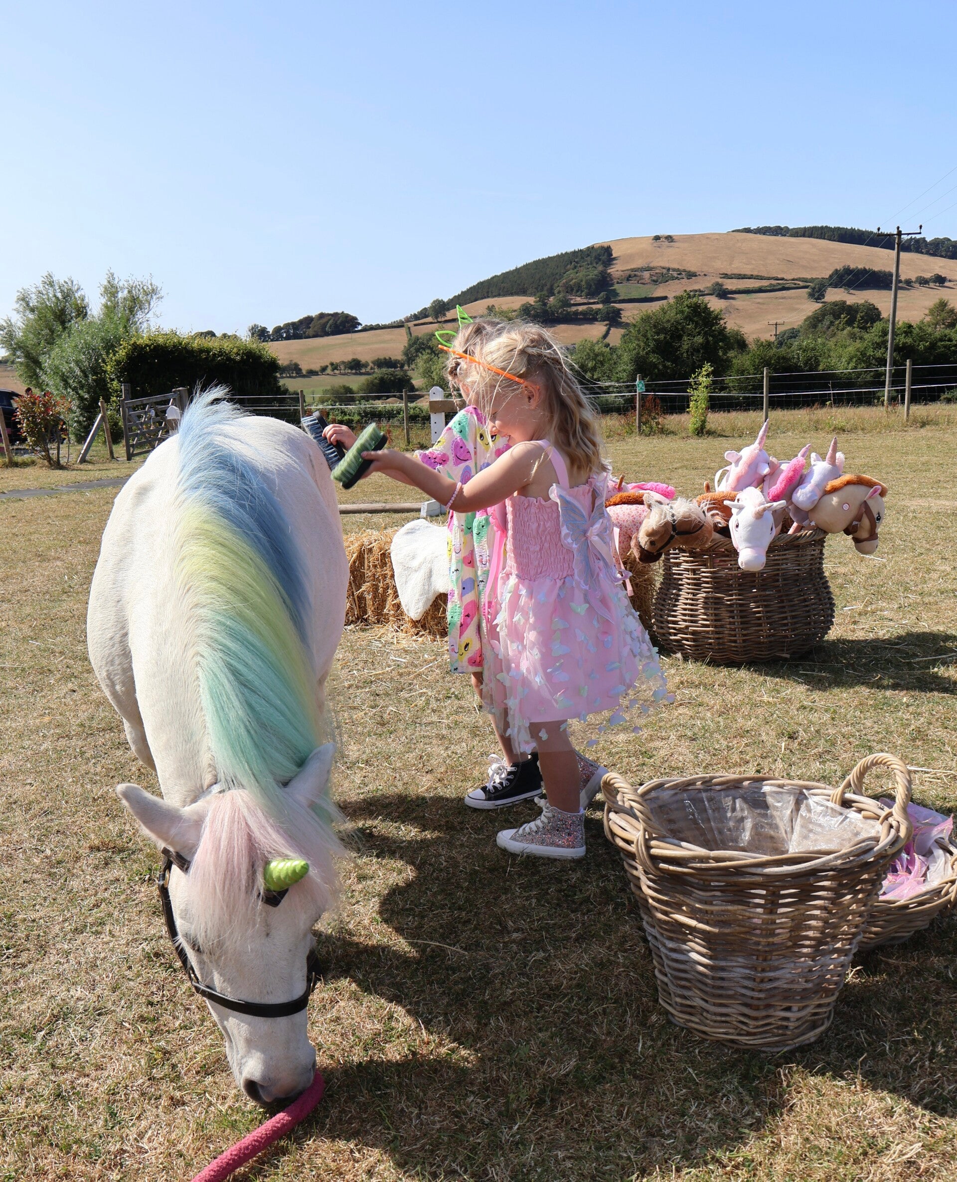 A happy child sat by the craft table with colouring books smiling at the white unicorn pony stood beside them. In the background there is a pastel coloured material backdrop with a neon 'let's celebrate' sign in the middle. 