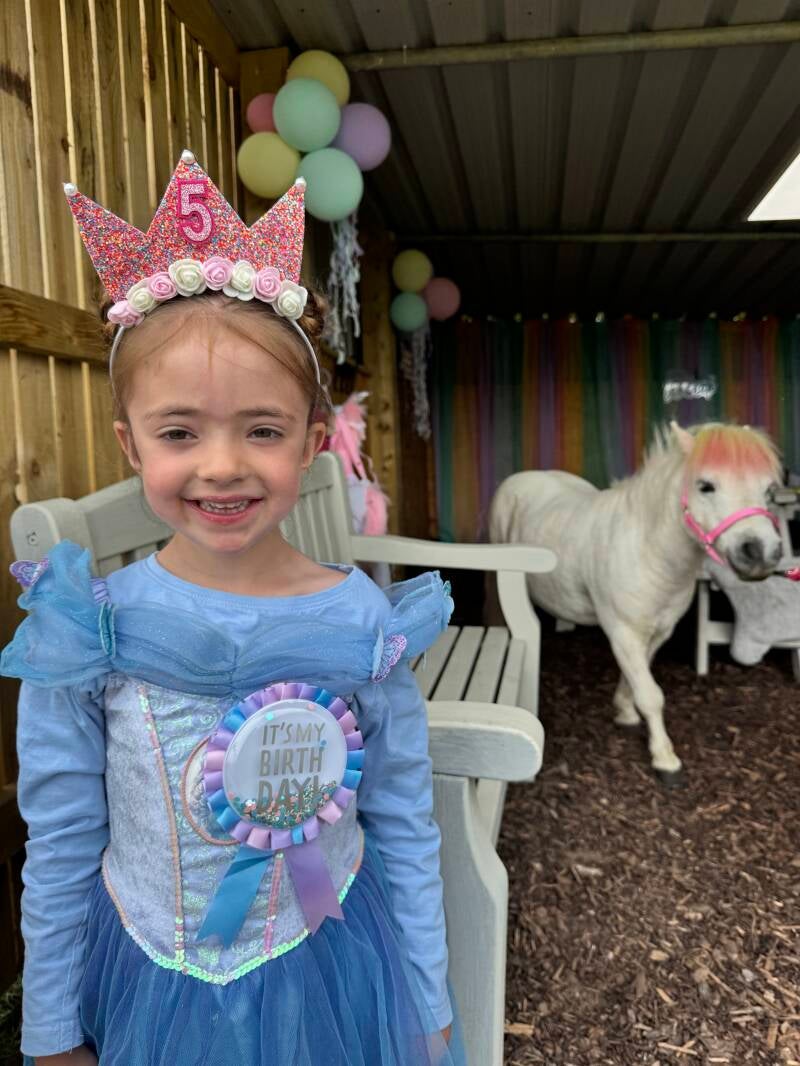 Child enjoying their 5th birthday party. Smiling at the camera with the unicorn pony and balloons in the background 