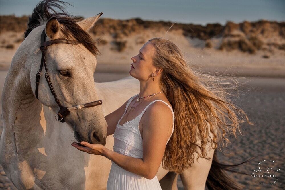 Junge Frau mit wehendem Haar vor Düne am Strand mit ihrem Pferd.