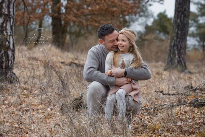 Papa kniet mit Tochter im Schoss im Gras und lacht.