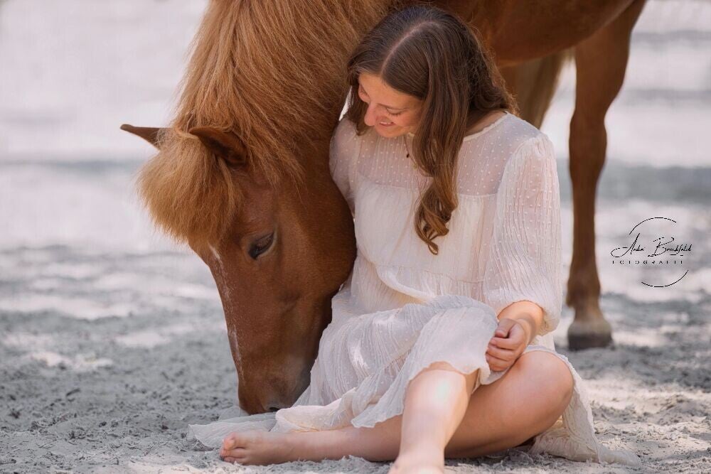 Junde Frau sitzt im Sand vor ihrem Pferd.