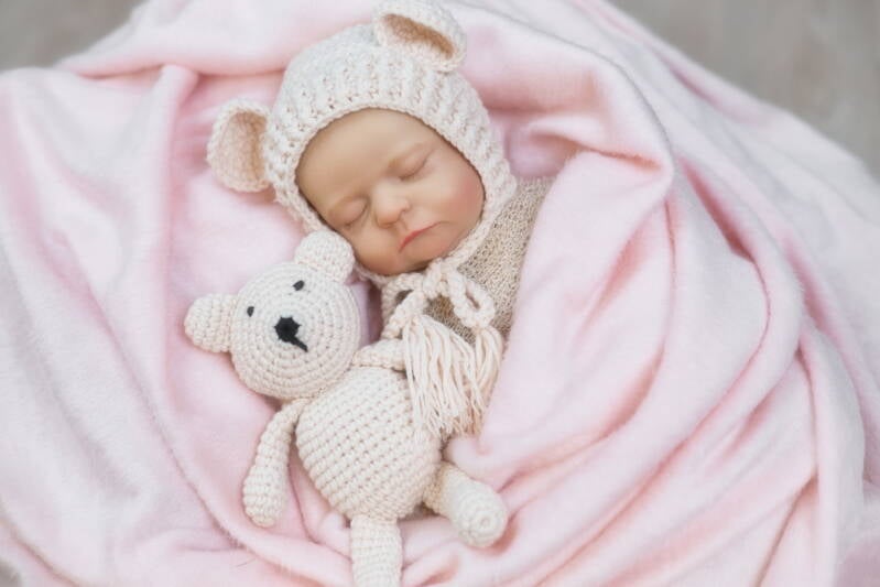 Neugeborenes mit Bärenmützchen schläft in Decke mit Bär im Arm.