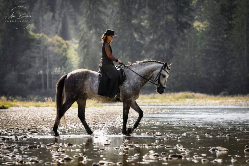 Frau reitet mit Reitrock und Zylinder durch einen Fluss.