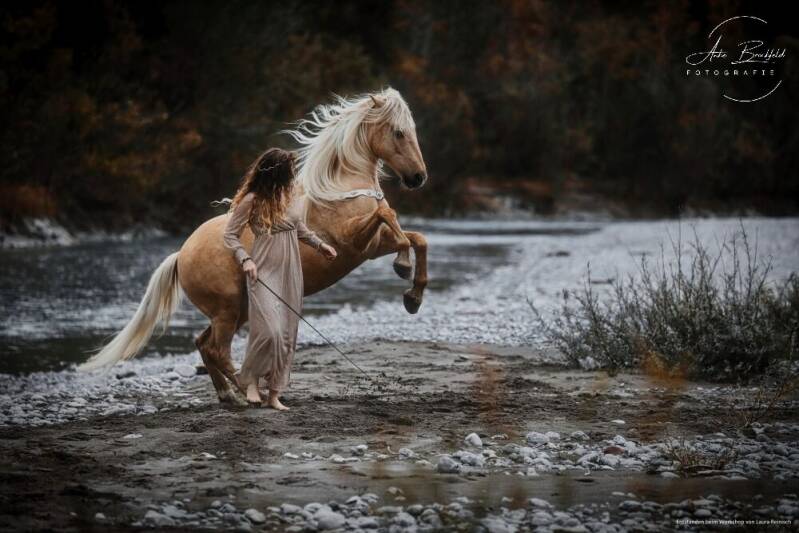 Frau im Kleid spielt frei mit Pferd am Fluss.