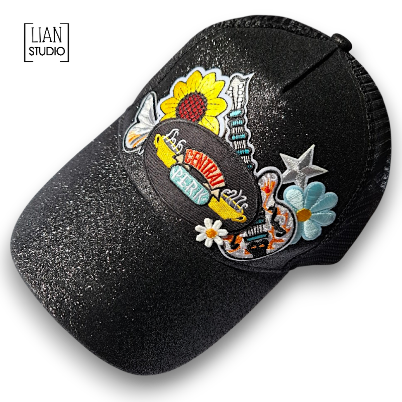 Zwarte glitter cap met mesh achterzijde een een groep strijk emblemen waarin de koffiebar van 'vrienden' centraal staat. Hier komen ze om alle belangrijke zaken met elkaar te bespreken. Phoebe neemt haar gitaar mee, met bloemen, vlinder en ster als geheel