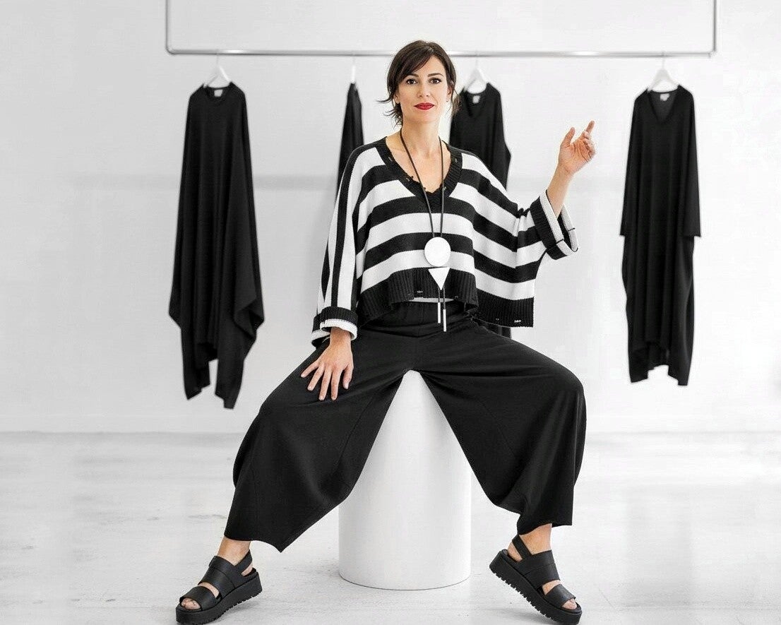 Eine modern gekleidete Frau sitzt auf einem weißen Hocker in einem eleganten Modegeschäft.  Die Frau trägt einen weiten,  schwarz-weißen Pulli und dazu eine weite schwarze Leinenhose, dazu kombiniert sie schwarze Sandalen mit dicker Sohle