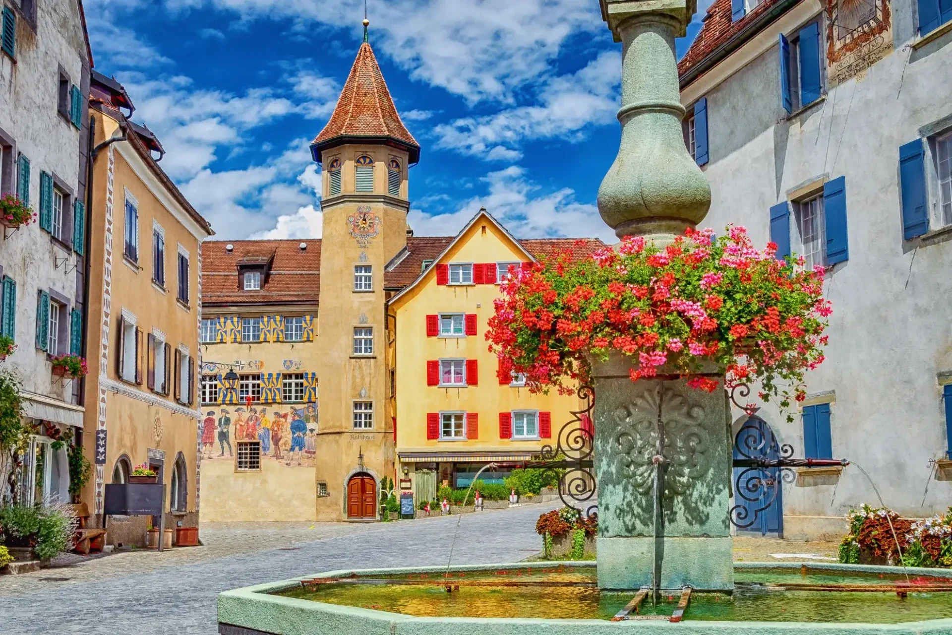 historischer-dorfplatz-in-maienfeld-mit-einem-blumengeschm-ckten-steinbrunnen-im-vordergrund-farbenfrohen-h-usern-mit-fensterl-den-und-dem-markanten-turm-von-schloss-salenegg-unter-blauem-himmel-standard.webp