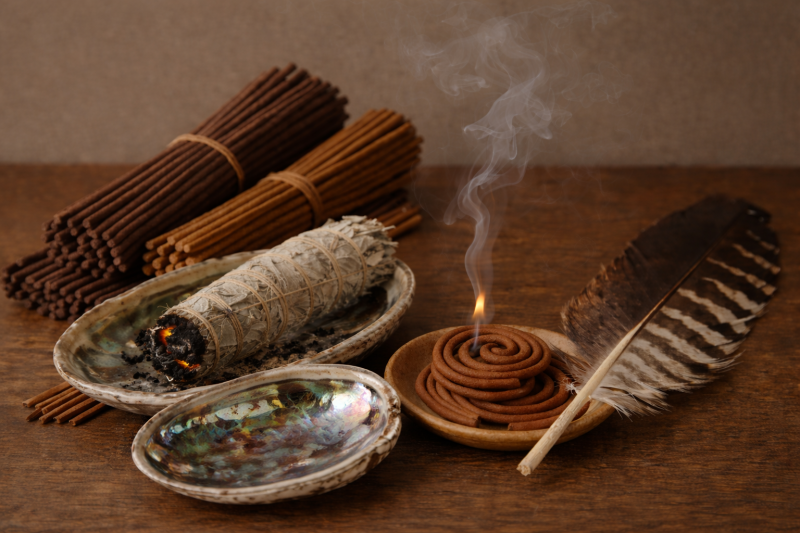 Stilleven van smudgingmaterialen op een houten tafel: bundels wierookstokjes, een brandende salie-smudge stick in een schelp, een spiraalvormige wierookkegel die rook afgeeft, een grote veer en twee glanzende abalone schelpen met iriserende kleuren.