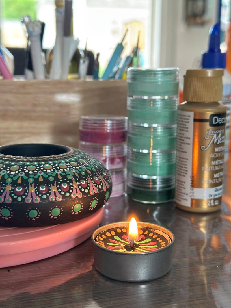 Handgemaakte mandala waxinelichthouder met kleurrijke stipkunst naast een brandend kaarsje en dot painting tools op creatieve werkplek.