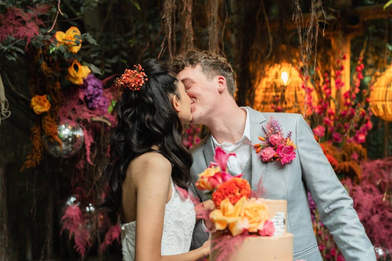 Een bruiloft in de Jungledome Centerparcs Heijderbos. Bruidspaar kust elkaar na het aansnijden van de bruidtaart. Een prachtige backdrop van kleurrijke bloemen op de achtergrond. Anders trouwen.