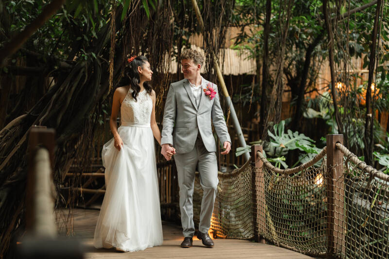 Een bruidspaar loopt hand in hand over een bruggetje en kijkt elkaar verliefd aan. De Jungledome van Centerparcs Heijderbos is een mooie  trouwlocatie in Nederland waar het altijd mooi weer is, ongeacht het seizoen. Tropische bruiloft.