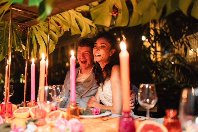 Bruidspaar zit aan de dinertafel in Jungle Dome Center Parcs Heijderbos. Kleurrijke kaarsen, bloemen, citrusvruchten en tropische bladeren. Prachtig! Wedding and Dreams. Leafsum. Janneke design.