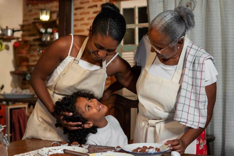 African nanny and elderly caregiver in aprons warmly engaging with a smiling child in a cozy kitchen, preparing food together and showing nurturing, family-centered care.