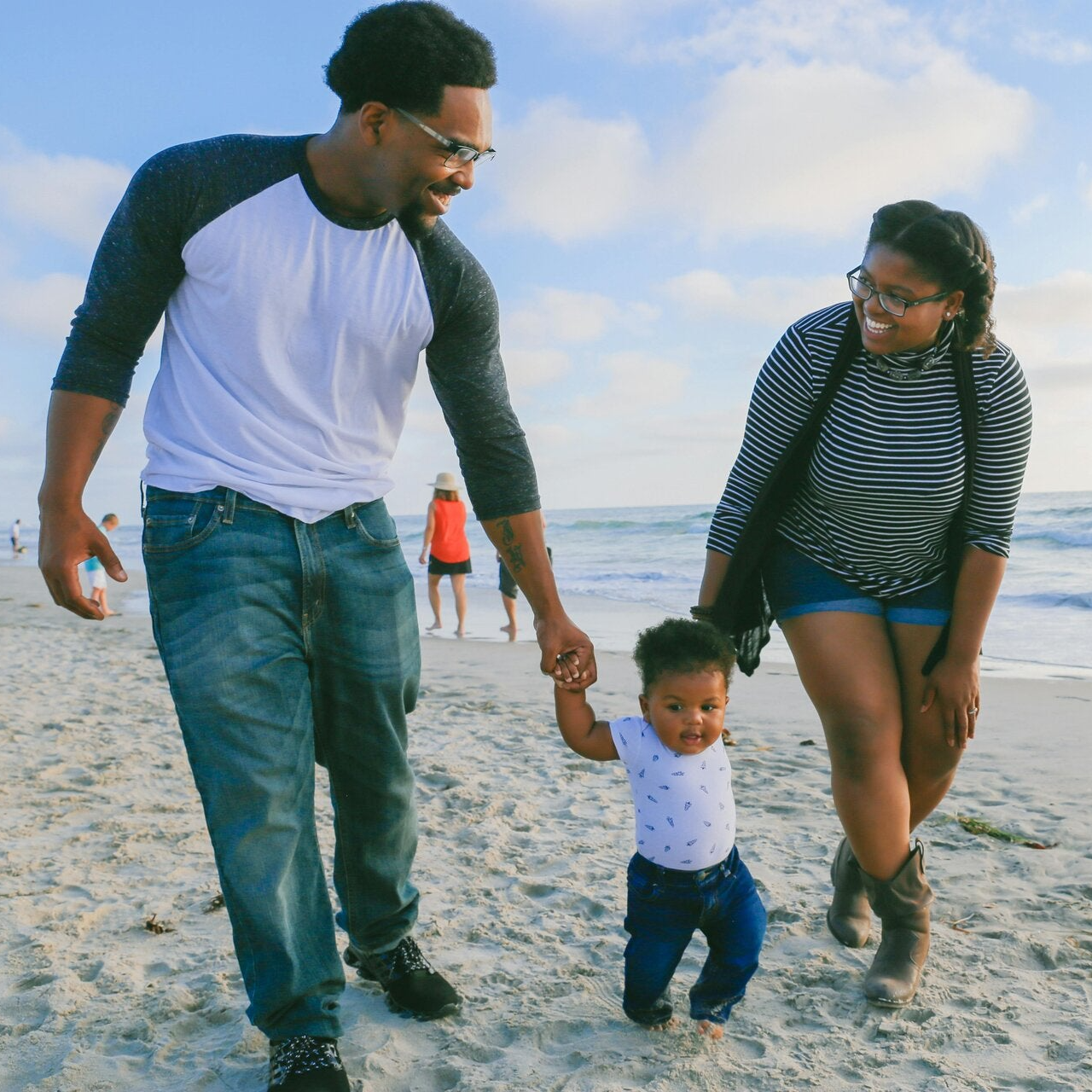 A young Black couple walks hand-in-hand along the beach with their toddler between them, symbolizing love, unity, and family protection in a peaceful, natural setting.