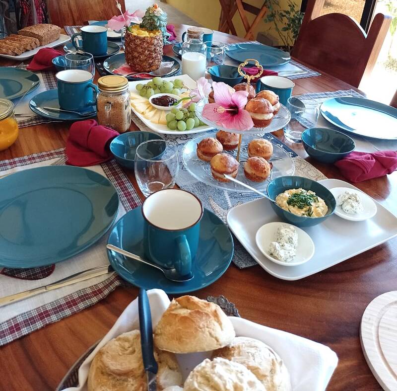 "Ein liebevoll gedeckter Frühstückstisch in Costa Rica mit frischen deutschen Semmeln, selbst gebackenem Brot, Muffins, Dips, tropischen Früchten und Hibiskusblüten als Dekoration – ein Ort, an dem deutsche Tradition und costa-ricanische Herzlichkeit mite
