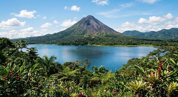 Majestätischer Vulkan Arenal in perfekter Kegelform hinter dem tiefblauen Arenalsee, umrahmt von üppiger grüner Tropen-Vegetation in Costa Rica.