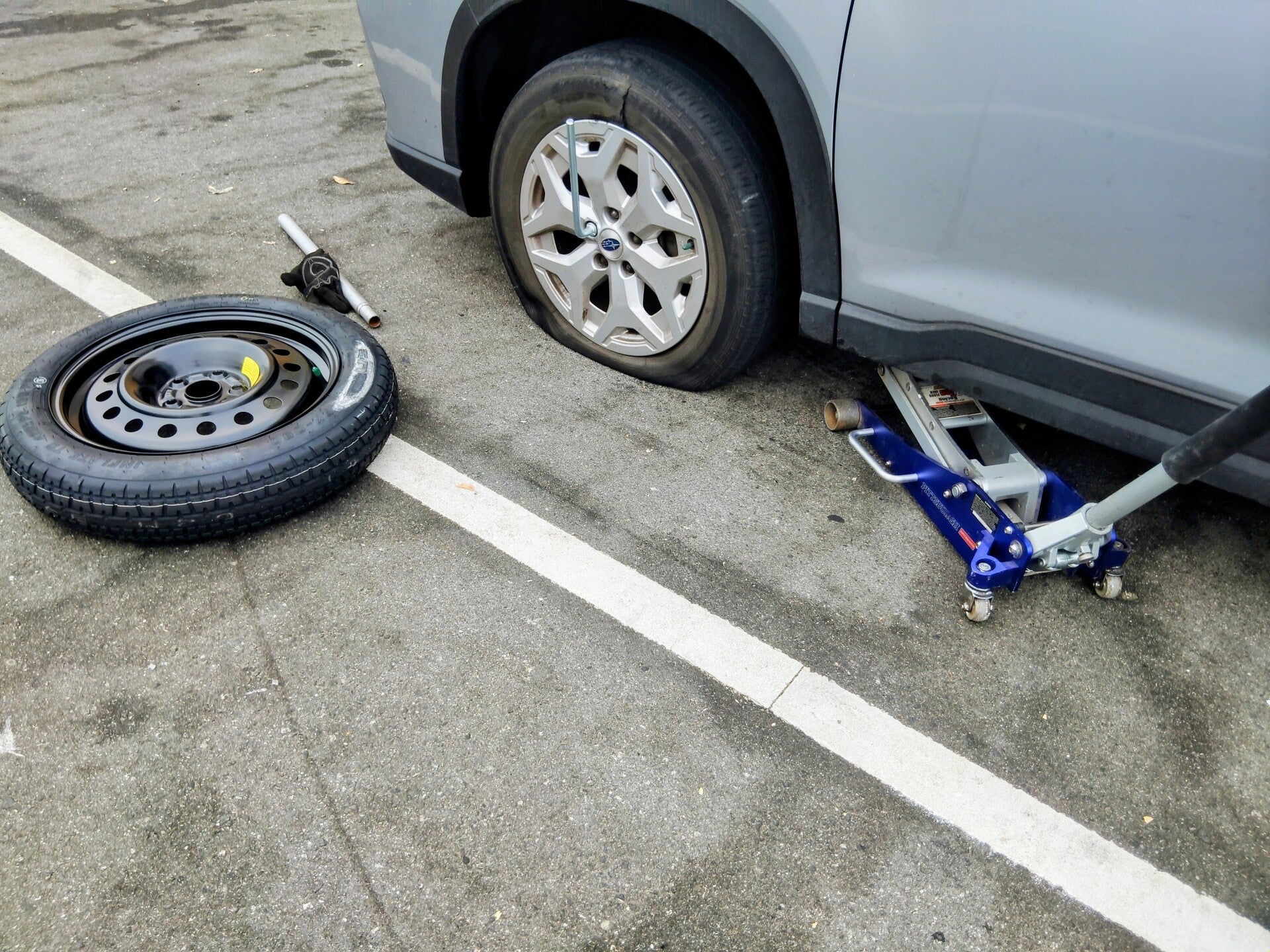 Silver Subaru Forester in the SF Bay Area with a flat tire being lifted by a blue hydraulic jack next to a spare tire.