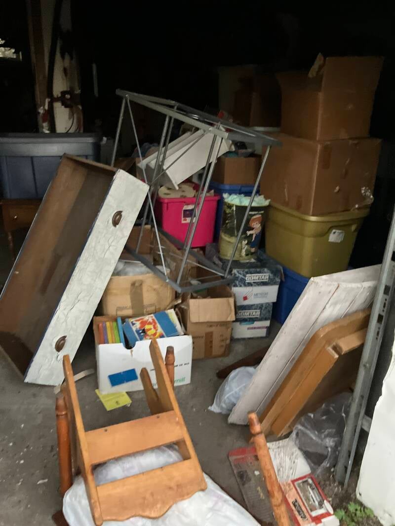 Cluttered storage area with boxes, furniture, and household belongings before cleanout.