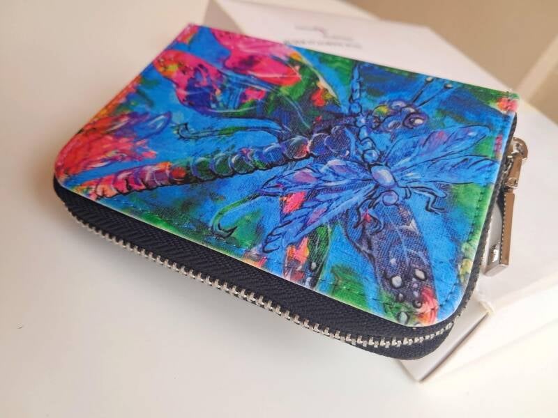 A compact 12cm × 9cm secure wallet featuring dragonfly artwork, full zip-around closure, interior card slots, and a coin pocket. Price 25€
