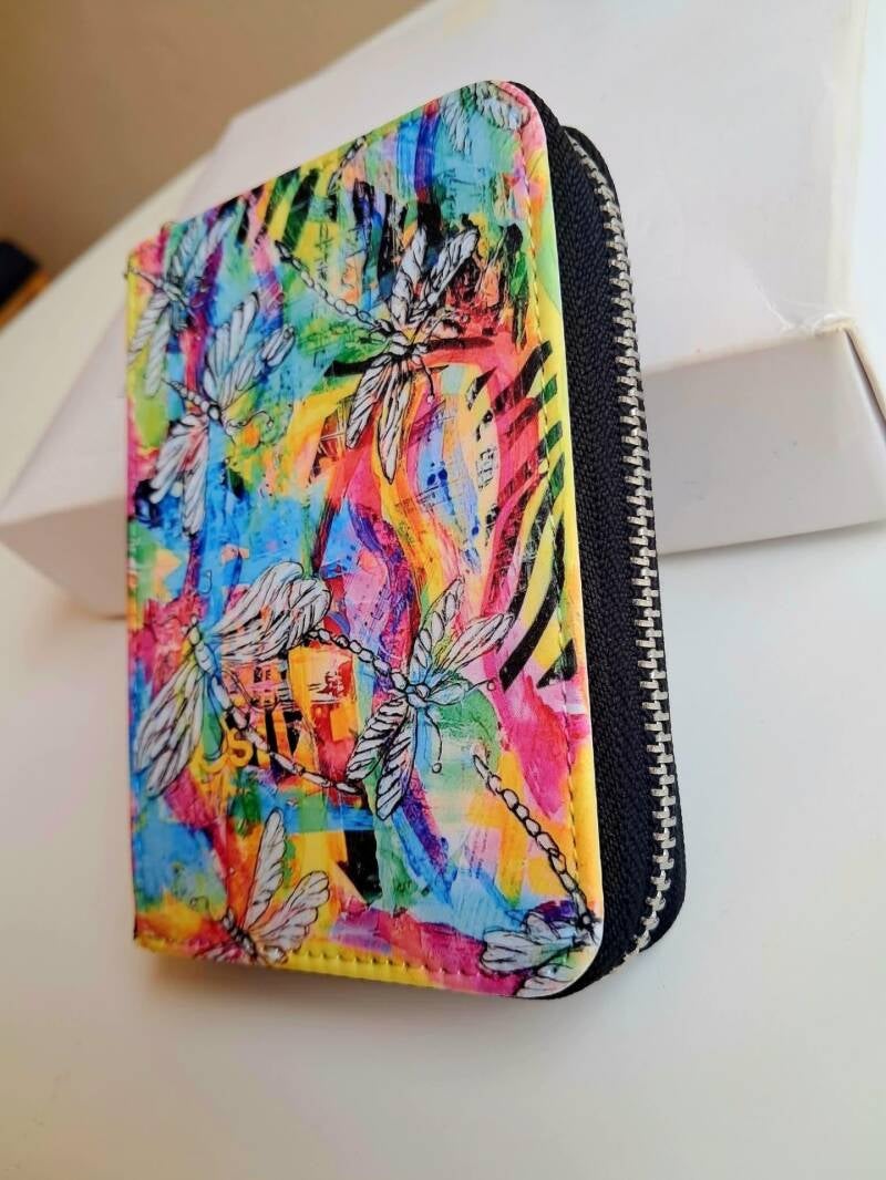 A compact 12cm × 9cm secure wallet featuring dragonfly artwork, full zip-around closure, interior card slots, and a coin pocket. Price 25€