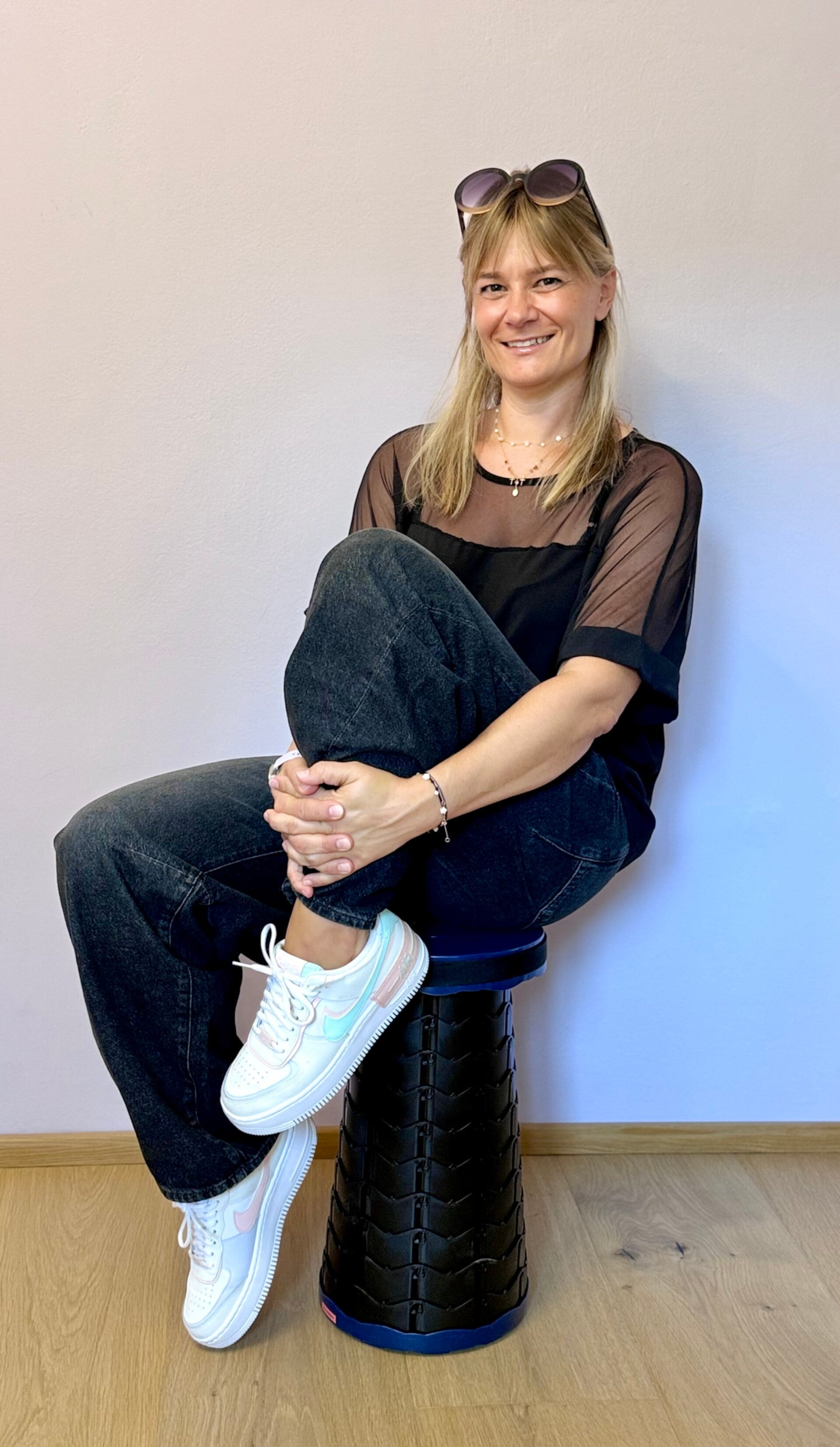 Jacqueline Pfanzelter sitzte vor hellilanen Wand auf Hocker, ist schwarz gekleidet, hat die Sonnenbrille in die Harre geschoben und lächelt