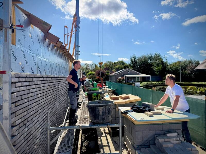 Gevel in opbouw met metselwerk, stelling en mensen op de werf.