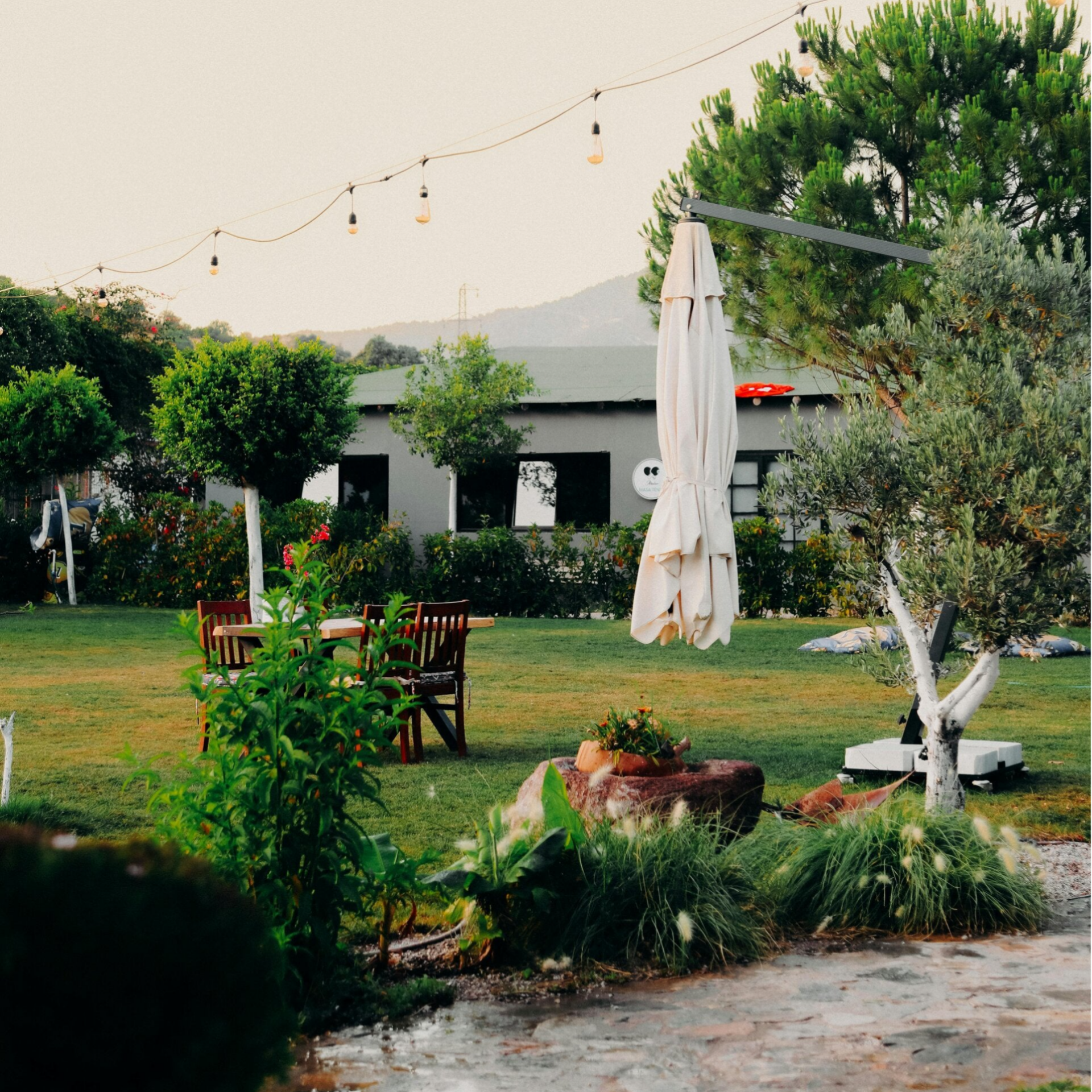 Das Bild zeigt einen gepflegten Garten, gemähter Rasen , einen Tisch mit Holzstühlen, einen geschlossenen Sonnenschirm. Im Hintergrund steht eine weie Villa..