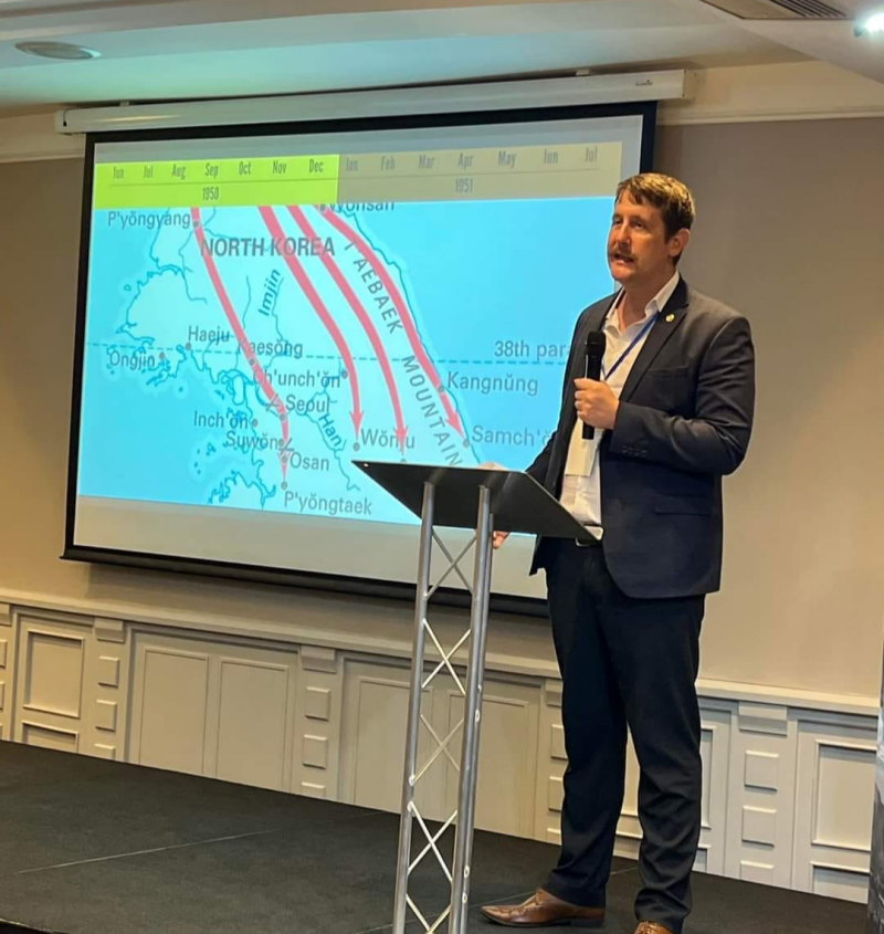 Historian delivering a presentation on the Korean War with a map of Korea displayed behind him.