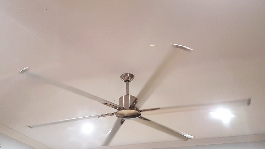 Large brushed-metal ceiling fan with long blades spinning, hung on a downrod from a white ceiling with LED downlights, installed by I.C.M