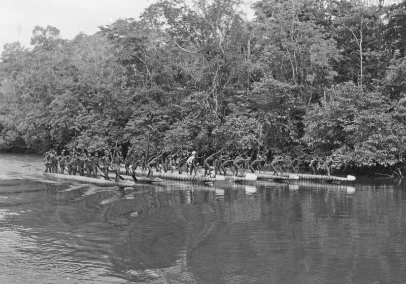 Bestuursambtenaren die met de tourneeboot Tasman een tocht maken door het Asmat-gebied zijn in de oorlogsprauwen van de Papoea's van de kampong Atsj overgestapt om een bezoek te brengen aan de kampong