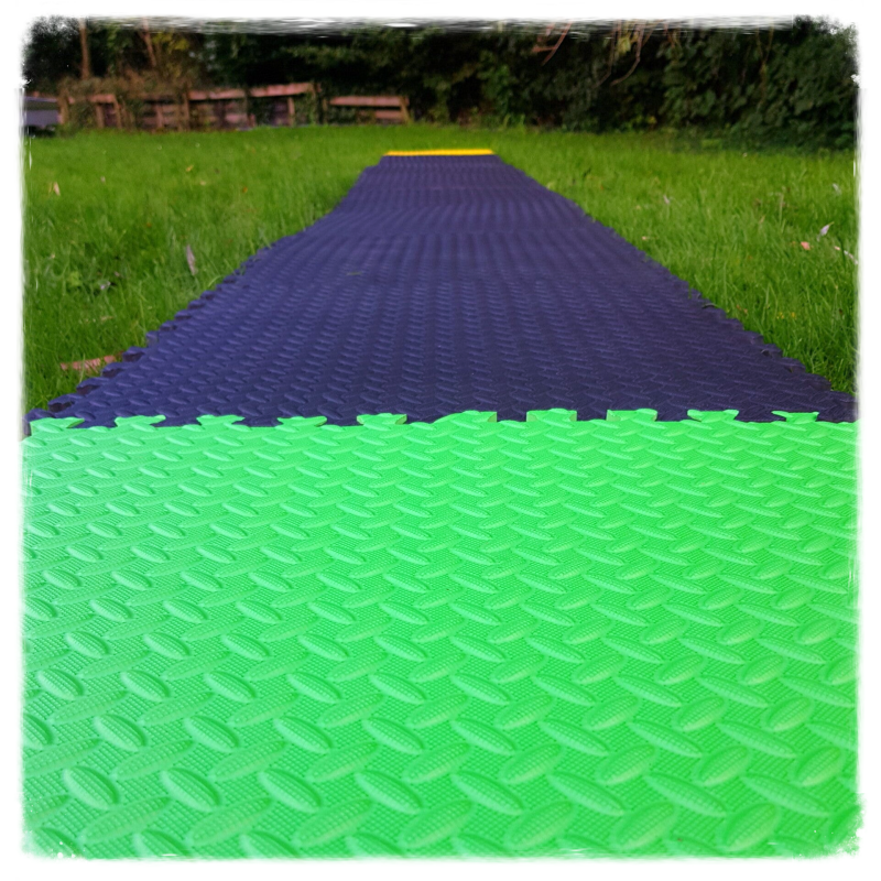 Een groene foam tegel ligt op de voorgrond, en via een reeks zwarte foam tegels gaat de route naar een gele foam tegel in de verte.  Het hoort bij de GridGame Run!, een serious game van GeckGenoeg.