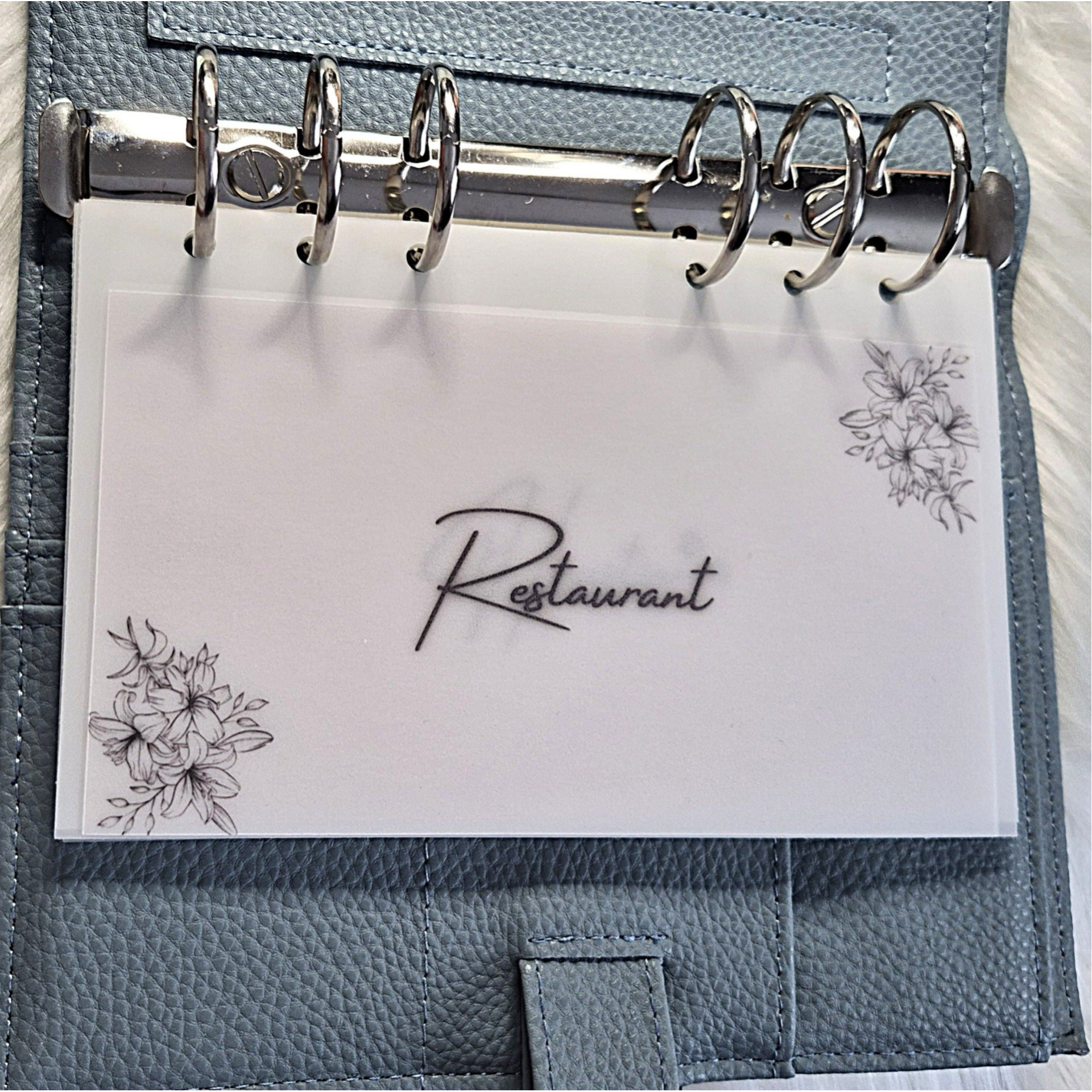Ein Produktfoto zeigt einen geöffneten, blaugrauen Budget Binder aus strukturiertem Leder mit einer silbernen 6-Ring-Mechanik. Darin abgeheftet ist ein personalisiertes, transparentes Dashboard als Geldumschlag für die Kategorie „Restau