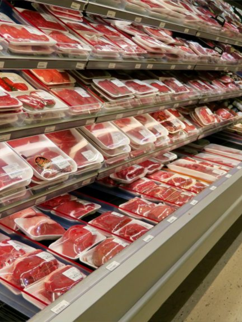 A grocery store meat aisle — refrigerated open cases filled with pre-wrapped cuts on white styrofoam trays, under overhead signs reading PORK and BEEF. The polished retail end of the supply chain. The crucifixion most people walk past every week and never see.