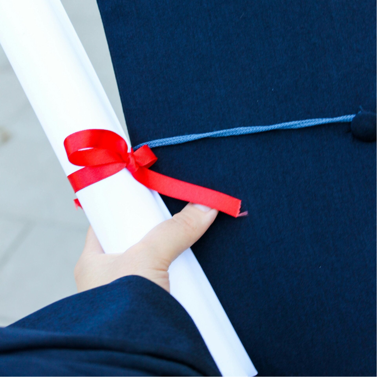 Hand holding a rolled diploma with red ribbon and a blue graduation cap with light blue tassel, representing my educational journey