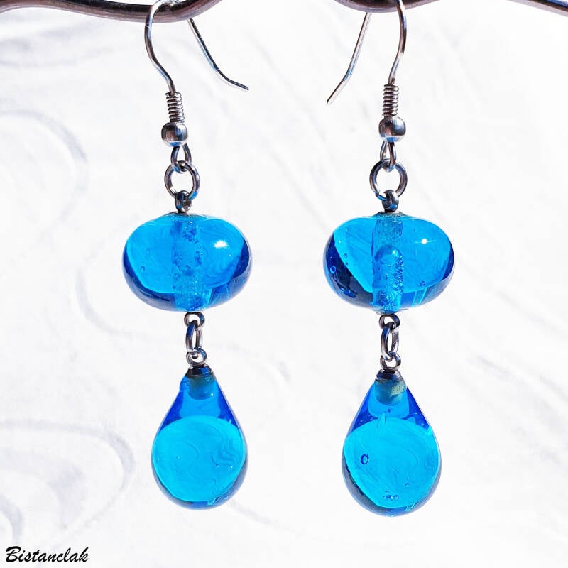 une paire de boucles d'oreilles en verre suspendue par leur crochet, elles sont composées chacune d'une perle en goutte surmontée d'une perle ronde de clouleur bleu turquoise transparent