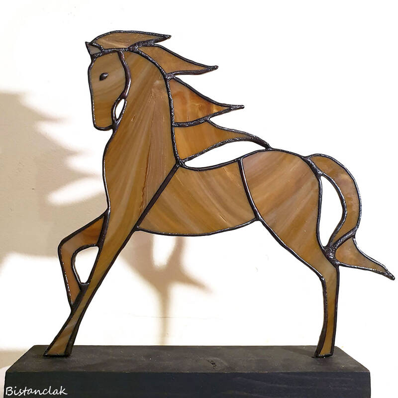 Cet objet de décoration vitrail représentant un cheval est exposé du côté où le verre coloré, dans des tons de sable, brun et beige, est plus brut et moins brillant, chaque face du cheval ayant une texture distincte.