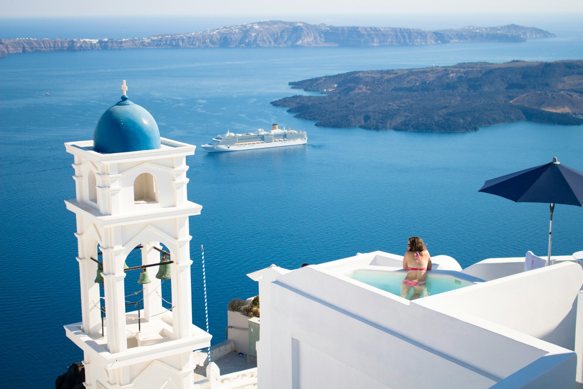 Luxury travel moment in Santorini: whitewashed cliffside views over the caldera with a cruise ship gliding across the deep-blue Aegean.
