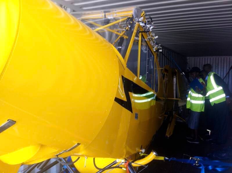 Kenyan customs staff in high-vis jackets inspecting a bright yellow Super Cub aircraft inside a shipping container, visibly confused—possibly by the plane, or the sneaky 200-litre avgas drum hidden among the ratchet straps and delusion.