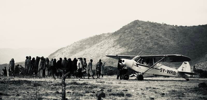 Piper Super Cub parked on a remote airstrip in Kenya beside a group of Maasai during Fly4Elephants operations. Silvertone field image showing conservation aviation, community presence, and bush flying in the Maasai Mara ecosystem.