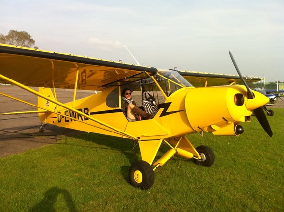 Nicole Romdane sits confidently in the cockpit of a bright yellow Piper Super Cub on a grassy airfield—still unaware that she has entered the danger zone of Campfire Syndicate’s “From Riches to Rags: An African Odyssey.”