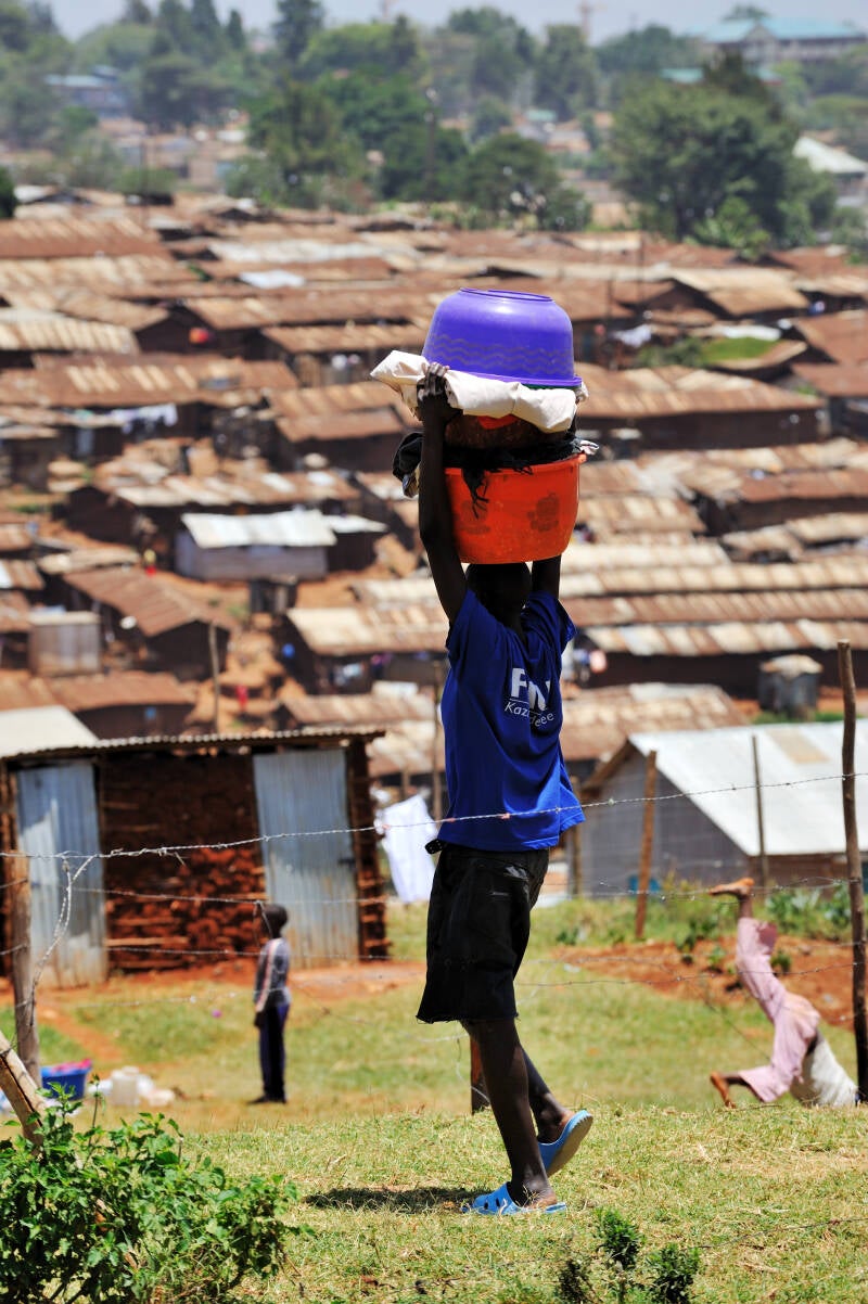 Boy in Kibera, Nairobi, carrying stacked plastic tubs on his head, standing before a sea of rusted tin rooftops — harsh sunlight, barbed wire, and daily survival, as captured in From Riches to Rags: An African Odyssey by Marcel Romdane.