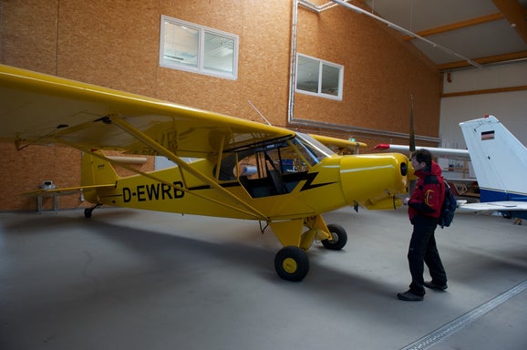 From Riches to Rags: An African Odyssey, Part VII – Love at First Stall. Flight instructor Enriko inspects the unsuspecting yellow Super Cub in Kalli’s hangar, seconds before signing up for a three-year descent into Romdane-fuelled madness.