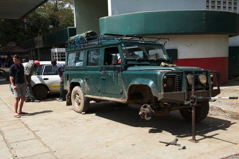 Land Rover Defender jacked up at a petrol station en route to Maasai Mara, Kenya, March 2012. Three flat tires, a leaking gearbox, and spark plug failure—documented in What Could Possibly Go Wrong?, a memoir of chaos and British mechanical betrayal.