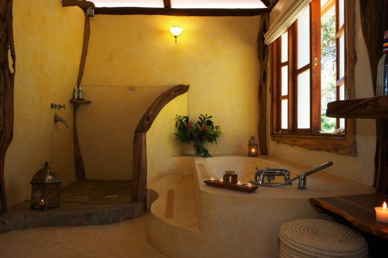 Luxury safari bathroom in Kenya with stone shower, hot bath, candles, and lemongrass soap—scene from What Could Possibly Go Wrong? by Romdane, where dark humour, collapse, and Campfire Syndicate rebellion meet five-star sedation.