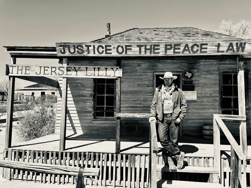 Marcel Romdane standing defiant at the Jersey Lilly Courthouse in Langtry, Texas — cowboy hat, boots, and outlaw posture included — blending frontier justice with Campfire Syndicate chaos in a monochrome moment that screams ‘Wanted: for Crimes Against Bei