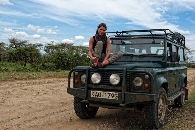 Nicole Romdane sits atop a green Land Rover Defender in rural Kenya. A scene from What Could Possibly Go Wrong?, this image captures safari chaos, post-colonial relics, and the calm before the mechanical storm of British engineering failure.