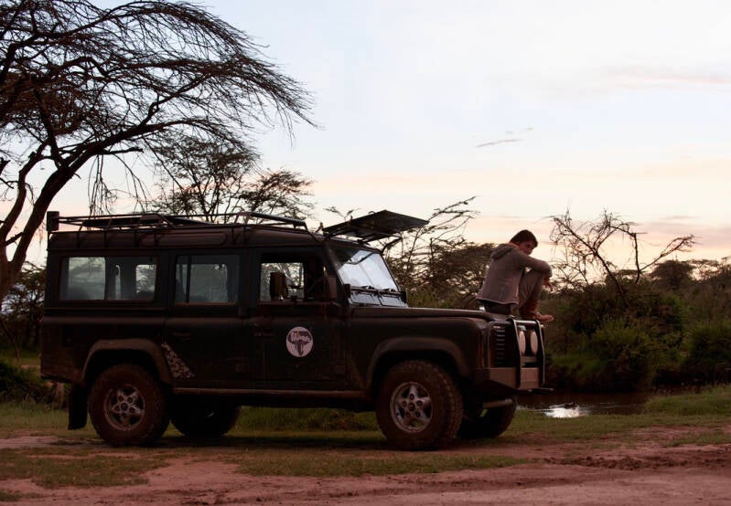 Nicole Romdane  sitting on the hood of a Land Rover Defender in the Maasai Mara at dusk, moments before a pride of lions arrived. Calm before the carnivore storm in Marcel Romdane’s absurd African saga The Hunger Games, Mara Edition.