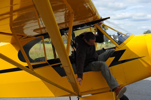 Marcel Romdane attempts (again) to enter a bright yellow Super Cub, gripping the frame while contorting mid-air. From Riches to Rags: An African Odyssey Part IX– Campfire Syndicate LLC.