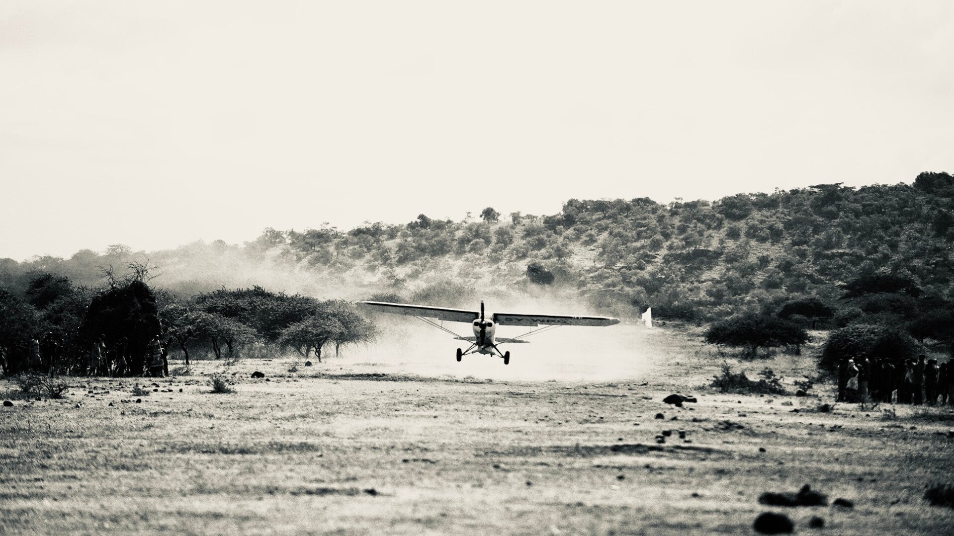 Piper Super Cub landing on a rough bush airstrip in Kenya during Fly4Elephants operations, raising dust as local observers stand along the strip. Field image representing conservation aviation, remote operations, and aerial support in East Africa.